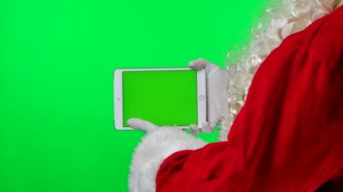 Beyaz eldivenli sakallı Noel Baba yeşil ekran krom anahtarı olan yatay pozisyonda tablet kullanıyor. Stüdyoda yeşil arka planda izole edilmiş. Ekranı düzelt. Kapatın. Yavaş çekim.