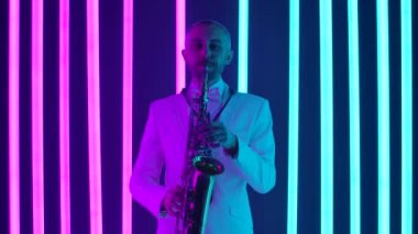 Beyaz takım elbiseli bir adam saksafonda beste yapıyor. Bir adam stüdyoda pembe ve mavi neon tüplerin arka planına karşı bir enstrüman çalıyor. Yaklaşın, ağır çekim..