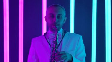 Beyaz takım elbiseli havalı bir saksafoncu parlak neon boruların arka planına karşı stüdyoda solo performans sergiliyor. Caz konsepti, retro müzik. Ağır çekim, yakın çekim..