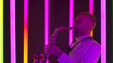 Beyaz takım elbiseli bir müzisyen ustaca altın saksafon çalıyor. Stüdyoda bir adamın yan görüntüsü çok renkli neon boruların arka planına karşı. Ağır çekim, yakın çekim..