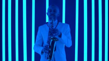 Caz saksafonu çalan deneyimli bir müzisyen. Beyaz takım elbiseli bir saksafoncu stüdyoda çok renkli neon ışıkların arka planına karşı solo performans sergiliyor. Müzik sanatı konsepti. Ağır çekim, yakın çekim..