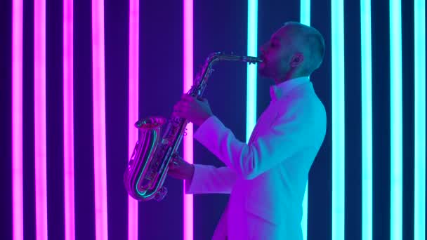 Vue latérale d'un jeune homme en costume blanc jouant magistralement du saxophone. Le musicien donne un concert en direct dans un studio sombre sur fond de néons lumineux. Ralenti, gros plan. — Video