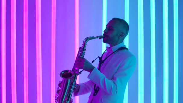 Vista lateral de un saxofonista en traje blanco tocando un instrumento musical. Un hombre toca música en un saxofón retro en un estudio oscuro con un telón de fondo de luces de neón rosadas y azules. En cámara lenta, de cerca. — Vídeos de Stock