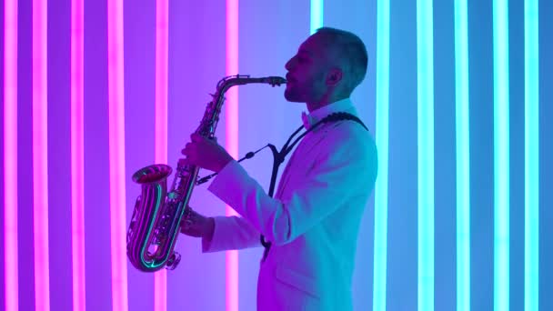 Vista lateral de un joven con un traje blanco tocando el saxofón magistralmente. El músico da un concierto en vivo en un estudio oscuro con el telón de fondo de luces de neón brillantes. En cámara lenta, de cerca. — Vídeo de stock