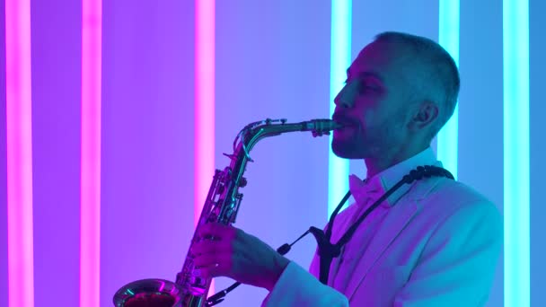 A man in a white suit plays a shiny gold saxophone. The dark studio is lit by bright pink and blue neon tubes. Neon light effects. Side view. Slow motion, close up. — Stock Video