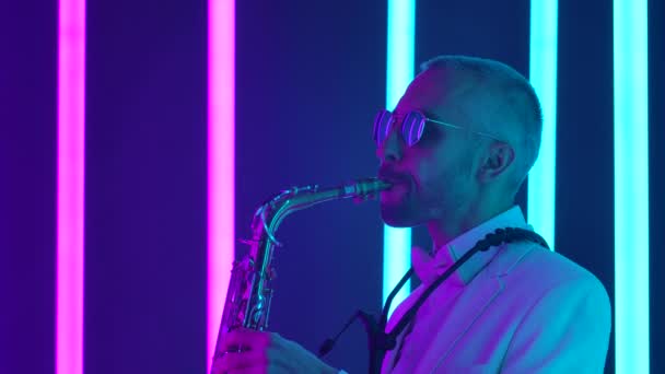 Un músico elegante con gafas de sol y un traje blanco toca el saxofón magistralmente. Un hombre actúa en vivo en un estudio con luces de neón brillantes. En cámara lenta. De cerca.. — Vídeo de stock