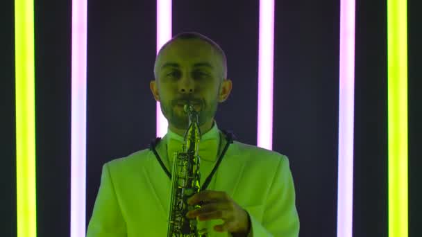 Um homem toca saxofone no estúdio contra o pano de fundo de tubos de néon multicoloridos. Um saxofonista em um terno branco realiza um solo incrível. Popularização do jazz. Movimento lento, close-up. — Vídeo de Stock