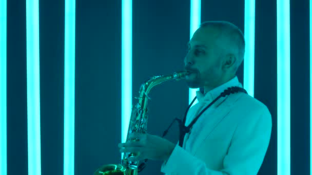 Un saxofonista profesional toca su instrumento. Un hombre con un traje blanco en un estudio oscuro iluminado por tubos de neón multicolores brillantes. Efectos de luz de neón. Vista lateral. En cámara lenta, de cerca. — Vídeos de Stock