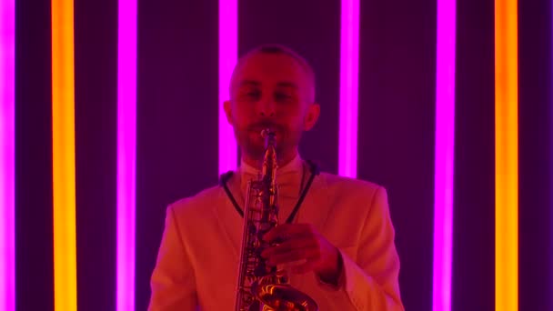 Un hombre con traje está realizando magistralmente una composición en el saxofón. El músico toca un instrumento musical en el estudio contra el fondo de tubos de neón multicolores. Primer plano, cámara lenta. — Vídeo de stock