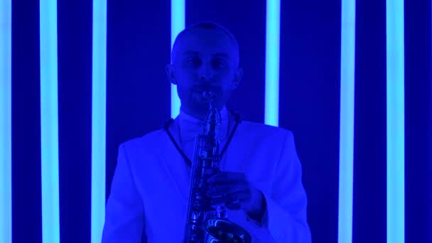 Utalentowany saksofonista gra jazz na złotym saksofonie. Mężczyzna w białym garniturze w studiu na tle kolorowych neonowych rur. Zwolnij, zamknij się.. — Wideo stockowe