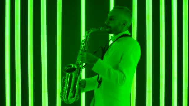 En professionell saxofonist spelar sitt instrument. En man i vit kostym i en mörk studio upplyst av ljusa flerfärgade neonrör. Neonljuseffekter. Sidovy. Långsam rörelse, närbild. — Stockvideo