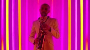 Beyaz takım elbiseli havalı bir saksafoncu, parlak renkli neon boruların arka planına karşı stüdyoda solo performans sergiliyor. Caz konsepti, retro müzik. Ağır çekim, yakın çekim..