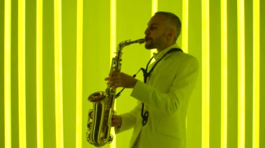 Beyaz takım elbiseli bir adam parlak altın saksafon çalar. Parlak renkli neon tüpleri karanlık stüdyoyu aydınlatıyor. Neon ışık efektleri. Yan görüş. Ağır çekim, yakın çekim..