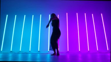 Çekici aktris mavi ve pembe neon tüplerin arka planında hassas şehvetli bir tefekküre daldı. Yetenekli genç bir dansçı ağır çekimde modern dans hareketleri sergiliyor. Siluet.