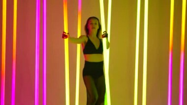 Bir dansçı, parlak renkli neon tüplerle çevrili bir stüdyoda modern bir dans sergiliyor. Elleri ağır çekimde neon tüplere tutunarak dans eden bir kız..