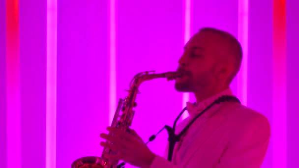 Profesionální saxofonista hraje na svůj nástroj. Muž v bílém obleku v tmavém studiu osvětleném pestrobarevnými neonovými trubicemi. Boční pohled. Živé vystoupení. Jazzová hudba. Zpomalit, zavřít. — Stock video