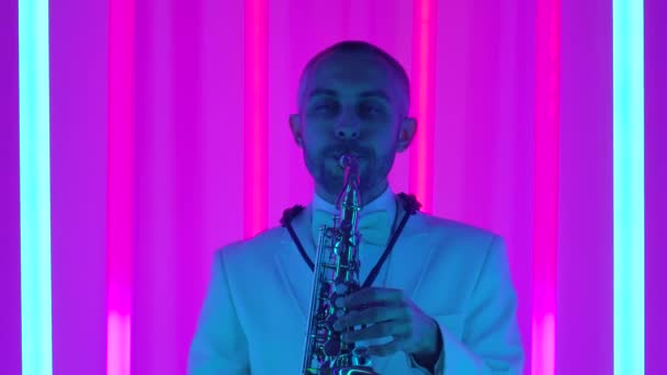 En man spelar saxofon i studion mot bakgrund av flerfärgade neonrör. En saxofonist i vit kostym utför ett fantastiskt solo. Popularisering av jazz. Långsam rörelse, närbild. — Stockvideo