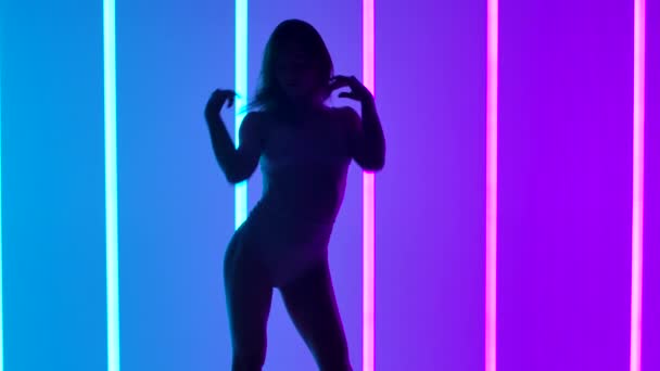 Suloisen naistaiteilijan siluetti, joka esittää nykykoreografian tanssielementtejä studiossa. Tyttö tanssii hidastettuna taustalla vaaleanpunainen ja sininen neon putket. Sulje se.. — kuvapankkivideo