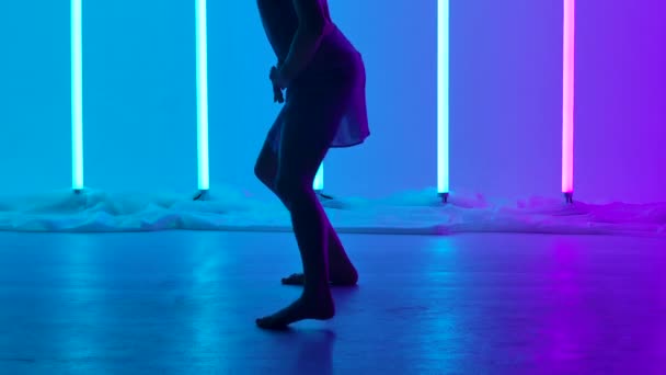O actriță desculță efectuează o dispreț blând în studio pe fundalul tuburilor de neon multicolore. O apropiere a picioarelor goale ale unui dansator calificat care face un lanț de pași de dans în mișcare lentă . — Videoclip de stoc