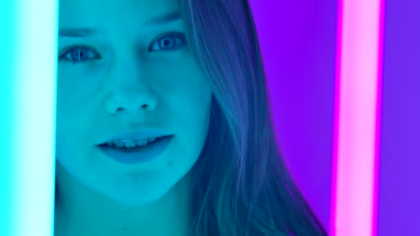 Retrato de una bailarina profesional caucásica. Primer plano de una cara de chicas sonrientes en un estudio oscuro cerca de tubos de neón brillantes. Una joven mujer se endereza y empuja su cabello hacia atrás en cámara lenta. — Vídeos de Stock