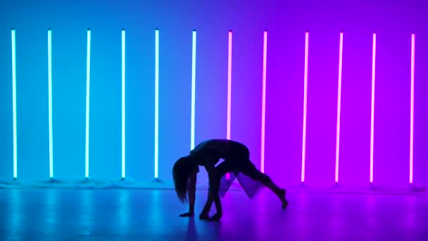 Een silhouet van een danseres die acrobatische elementen uit de hedendaagse choreografie opvoert. Een kunstenaar danst in een studio tegen een achtergrond van blauwe en roze neon buizen in slow motion. — Stockvideo