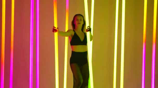 Een danser voert een moderne dans uit in een studio omringd door felgekleurde neonbuizen. Een meisje danst vasthouden aan neon buizen met haar handen in slow motion. — Stockvideo