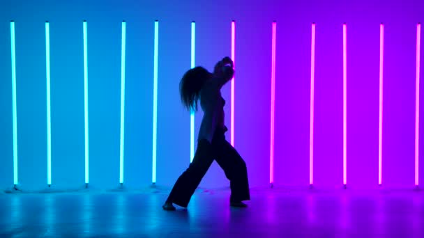 Fiatal nő táncos táncol kortárs a stúdióban hátterében sokszínű neon csövek. Egy tehetséges lány táncol a modernitás és a balett elemeivel. Sziluett. Lassú mozgás. — Stock videók