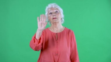 Yaşlı kadının portresi el sallıyor ve el sallıyor. Buraya gel. Stüdyoda yeşil ekranda kırmızı bluzlu gri saçlı büyükanne. Kapat..