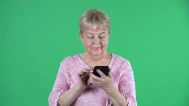 Akıllı telefonuyla mesaj atan yaşlı bir kadının portresi. Stüdyoda yeşil ekranda pembe kazaklı, kısa saçlı gri saçlı büyükanne. Kapat..