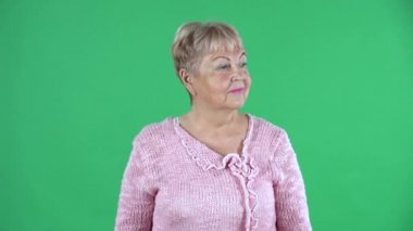 Yaşlı bir kadının portresi etrafa bakınıyor ve gizli bir jest yapıyor. Stüdyoda yeşil ekranda pembe kazaklı, kısa saçlı gri saçlı büyükanne. Kapat..