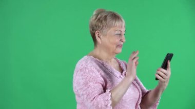 Yaşlı kadının portresi akıllı telefonuyla bir video görüşmesinde konuşuyor. Stüdyoda yeşil ekranda pembe kazaklı, kısa saçlı gri saçlı büyükanne. Yan görüş. Kapat..