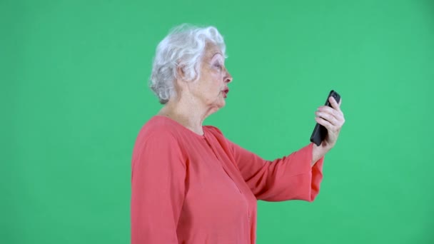 Portrait einer älteren Frau, die sich bei einem Videoanruf mit ihrem Smartphone unterhält. Grauhaarige Großmutter in roter Bluse auf grünem Bildschirm im Studio. Seitenansicht. Nahaufnahme. — Stockvideo