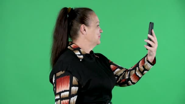 Retrato mulher idosa falando em uma chamada de vídeo usando seu smartphone. Morena avó em vestido preto na tela verde no estúdio. Vista lateral. Fechar. — Vídeo de Stock