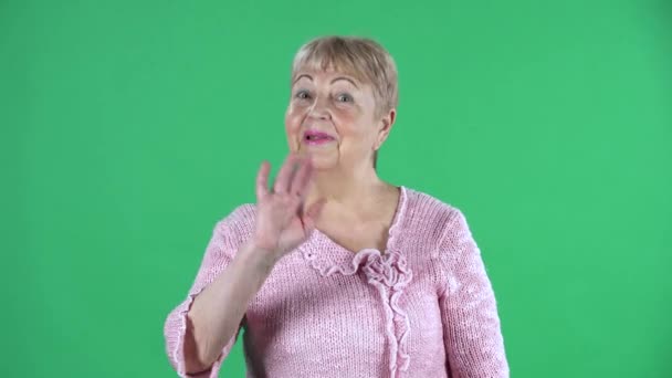 Portrait einer älteren Frau, die in die Kamera winkt und Gesten zeigt. Grauhaarige Großmutter mit kurzen Haaren im rosafarbenen Pullover auf grünem Bildschirm im Studio. Nahaufnahme. — Stockvideo