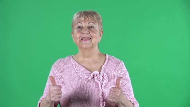 나이든 여성이 카메라를 보고 엄지손가락으로 제스 쳐를 보여 주고 있습니다. 회색 머리의 할머니가 스튜디오 녹색 화면에 핑크 색 스웨터를 입고 있습니다. 닫아. — 비디오