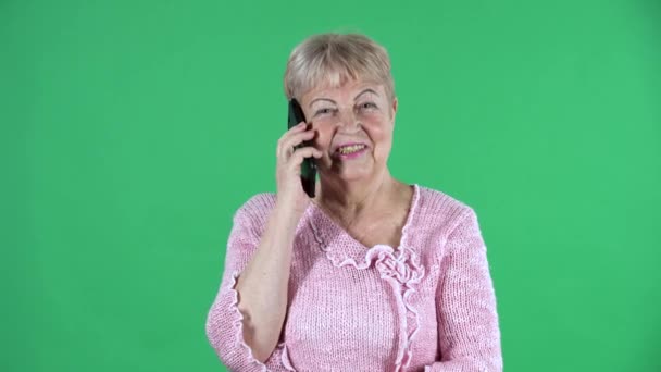 나이든 여성이 카메라를 보고 스마트폰으로 이야기하는 모습. 회색 머리의 할머니가 스튜디오 녹색 화면에 핑크 색 스웨터를 입고 있습니다. 닫아. — 비디오