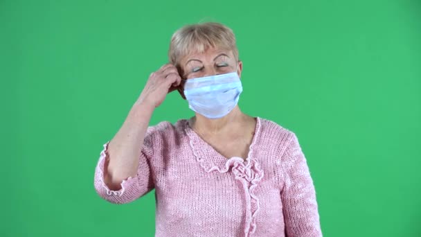 Retrato mujer anciana mirando a la cámara y quitándose la máscara médica. Abuela de pelo gris con el pelo corto en un suéter rosa en la pantalla verde en el estudio. De cerca.. — Vídeos de Stock