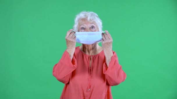 Retrato mulher idosa olhando para a câmera e rputs em máscara médica. Avó de cabelos grisalhos em blusa vermelha na tela verde no estúdio. Fechar. — Vídeo de Stock