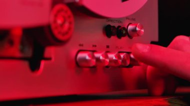 Erkek elleri, klasik analog analog makaradan makaraya, manyetik ses kayıt cihazını açar ve kapatır. Kırmızı neon ışıklarıyla aydınlatılan eski kayıt cihazının detaylı görüntüleri. Kapat..