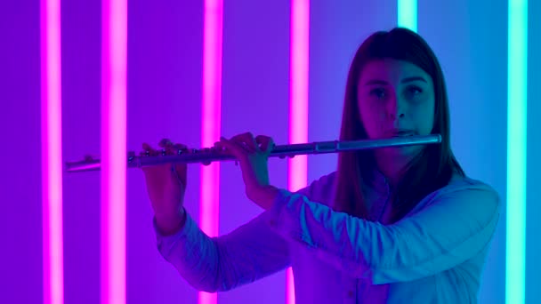 Cute young female musician plays the flute. Flute player professionally plays the wind instrument in dark studio against the backdrop of bright neon lights. Silhouette. Slow motion. Close up. — Stock Video