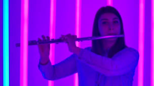 Potret musisi wanita muda bertiup ke dalam seruling instrumen angin. Seorang wanita flutist melakukan di studio gelap terhadap latar belakang lampu neon terang. Siluet. Gerakan lambat. Tutup.. — Stok Video