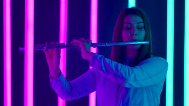 Un concert muzical de muzică clasică interpretat de o flutistă profesionistă. O tânără cântă muzică într-un studio întunecat pe fundalul luminilor luminoase de neon. Silueta. De aproape. Slow motion . — Videoclip de stoc