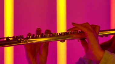 Müzikal tiyatro orkestrasında profesyonel flütçünün usta performansı. Elinde flüt olan bir kadın eli. Parlak renkli neon ışıkların arka planına karşı rüzgar enstrümanı. Yavaş çekim.