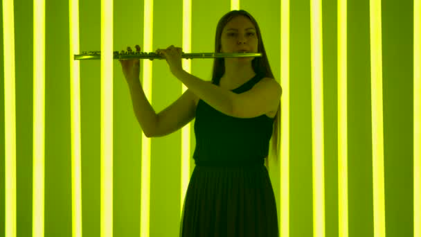 Flutist feminin fermecător într-o rochie neagră pe un fundal de lumini luminoase de neon într-un studio întunecat. Silueta unei tinere cântând o melodie la flaut. Slow motion . — Videoclip de stoc