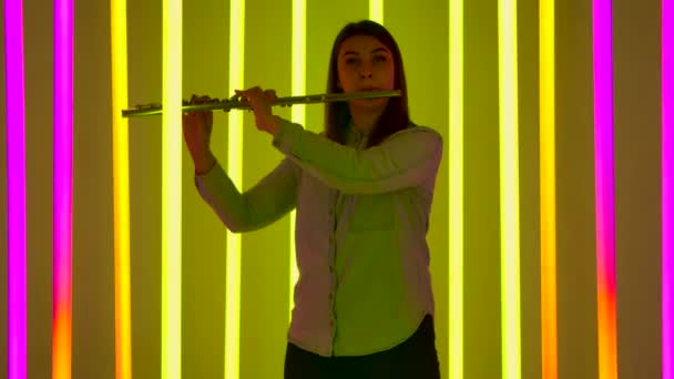 Retrato de jovem músico soprando na flauta do instrumento de sopro. Uma flautista feminina se apresenta em um estúdio escuro contra o pano de fundo de luzes de néon brilhantes. Silhueta. Movimento lento. — Vídeo de Stock