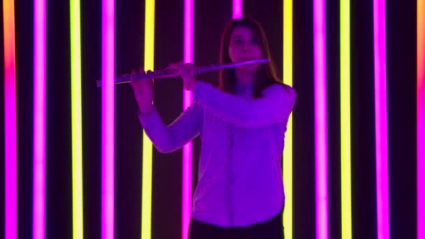 Een muzikaal concert van klassieke muziek uitgevoerd door een professionele vrouwelijke fluitist. Jonge vrouw speelt muziek in een donkere studio tegen de achtergrond van fel neonlicht. Silhouet. Langzame beweging. — Stockvideo