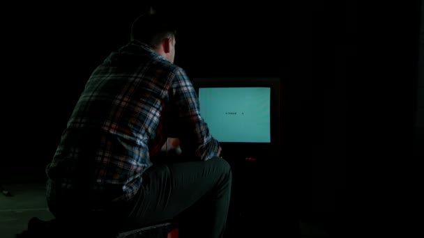 Vista trasera de un hombre jugando en una consola de juegos con un joystick mientras está sentado en una habitación oscura frente a un viejo televisor. Juego del legendario videojuego Tank. Videojuego retro para Nintendo de los 80. — Vídeos de Stock