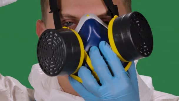 Portrait of a male medical doctor in a white protective suit and surgical gloves looking into the camera puts on a medical respirator. Green screen, chroma key. Slow motion. — Stock Video