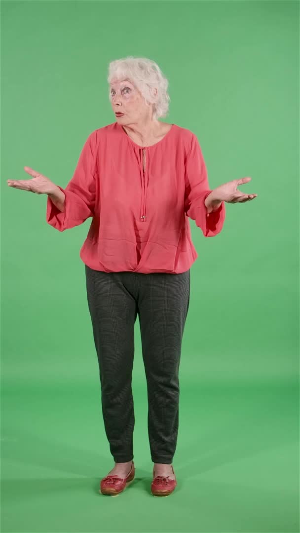 O femeie în vârstă care se uită la camera care-i scutură capul nu sunt eu, nu ştiu ce se întâmplă. Bunica cu părul gri în bluză roșie pe ecran verde la studio. Mişcare lentă. Video vertical . — Videoclip de stoc