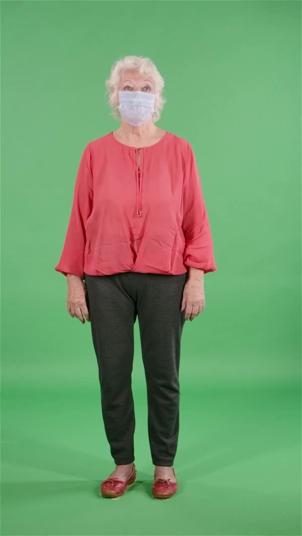 Портрет пожилой женщины, смотрящей в камеру и снимающей медицинскую маску. Серая бабушка в красной блузке на зеленом экране в студии. Медленное движение. Вертикальное видео. — стоковое видео